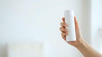 Woman's hand holding blank white cylindrical product container against neutral background for branding and packaging mockup purposes.