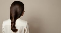 Young woman with long brown braided hair wearing white shirt viewed from behind against neutral beige background for beauty and haircare concepts.