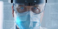 Healthcare professional in surgical mask and protective goggles examining digital medical interface with focused expression, representing modern medical technology in clinical setting.