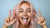 Young Caucasian woman with pink hair showing peace signs and sticking tongue out against blue background, expressing joy and playful attitude in studio portrait.