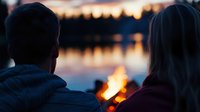 Couple enjoying romantic campfire by lake at sunset with warm golden light reflecting on water creating peaceful outdoor evening atmosphere.