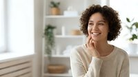 Young woman with curly dark hair and medium skin tone wearing cream sweater, smiling while touching face in bright modern interior with plants and shelving, suitable for lifestyle, wellness, and beauty marketing.