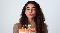 Young woman with long wavy brown hair blowing out colorful birthday candles on decorated cake, celebrating special occasion with joy and happiness.