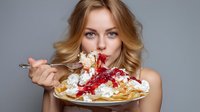 Young blonde woman with blue eyes eating dessert with whipped cream, strawberry sauce, and ice cream on crepes against gray background.