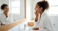 Young African American woman applying skincare cream in modern bathroom mirror reflection. Daily beauty routine and self-care wellness concept.