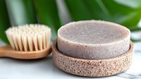 Natural solid shampoo bar with bamboo brush and cork holder on white marble surface against green leaves background, zero waste bathroom essentials.