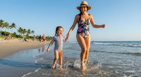 Mother and daughter running together on tropical beach during summer vacation, holding hands while splashing in ocean waves at sunset.