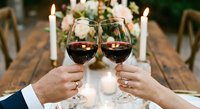 Elegant couple toasting with red wine glasses at romantic candlelit dinner celebration with white roses and candles creating intimate dining atmosphere.