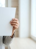 Person holding blank white paper sign with hand visible, arm extended forward in bright indoor space with blurred background for advertising and marketing campaigns.