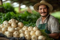 Hispanic farmer with mustache wearing straw hat and apron smiles while displaying fresh harvested white potatoes at local organic farm market, surrounded by produce.