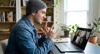 Happy young Caucasian man in gray beanie waving during video call on laptop at home office. Remote work communication and online meeting concept.
