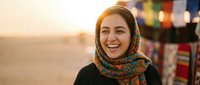Young Middle Eastern woman wearing colorful headscarf smiling joyfully at camera with warm sunset lighting and blurred market background for lifestyle content.