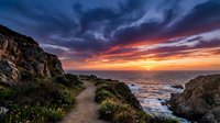 Dramatic sunset over rocky coastal landscape with golden and purple sky, ocean waves, and natural vegetation along cliff pathway.