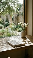 Peaceful morning scene with white flowers in ceramic vase on wooden table by window overlooking lush garden courtyard with natural sunlight and shadows.
