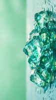 Crushed emerald green glass shards with water droplets on mint background, suitable for luxury cosmetics, jewelry, and wellness product packaging designs.