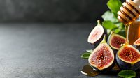 Fresh ripe figs cut in half with honey dripping from wooden dipper, green leaves on dark slate background with copy space. Food photography styling.