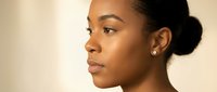 Young African American woman in profile with elegant low bun hairstyle and pearl earrings against neutral background for beauty and skincare concepts.