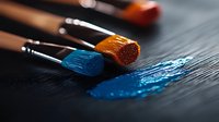 Paint brushes with vibrant blue and orange bristles resting on dark surface with fresh paint strokes, showing artistic tools in dramatic lighting and shallow depth of field.