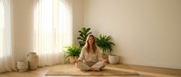 Young blonde woman practicing meditation in minimalist room with plants and natural light creating peaceful wellness atmosphere for mindfulness and relaxation.