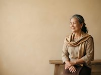 Senior Asian woman with long gray hair and glasses, wearing a beige cowl neck sweater, sitting on wooden bench against neutral background. Portrait for lifestyle and wellness content.