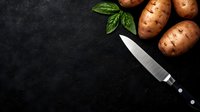 Fresh raw potatoes with green basil leaves and professional chef knife on dark black stone background with copy space for menu or recipe.
