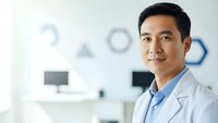 Professional Asian male doctor in white coat smiling confidently in modern medical office with computer workstation and clinical equipment background.