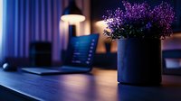 Purple lavender flowers in black ceramic pot on wooden desk with laptop and lamp in dark moody home office setting, creating peaceful work atmosphere.
