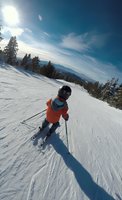 Young skier in bright orange jacket descending snowy mountain slope with panoramic winter landscape view and dramatic blue sky.