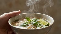 Hot steaming bowl of Asian noodle soup with fresh basil, lime wedge, and broth. Hand holding ceramic bowl. Use for food, cuisine, cooking, restaurant, and culinary content.