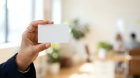 Young professional man with medium skin tone displaying blank white business card in modern office environment with colleagues and plants in background.