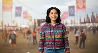 Young woman with dark hair and South Asian ethnicity wearing a colorful patterned zip-up jacket at an outdoor festival or event with crowds and decorative flags in background.