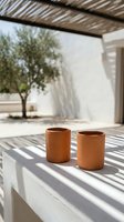 Two terracotta ceramic cups on concrete ledge under pergola with dappled sunlight shadows creating striped pattern in minimalist outdoor setting.