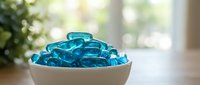 Blue gel capsules or supplements in white ceramic bowl on wooden table with natural lighting and green plant background for health and wellness concepts.