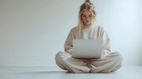 Young blonde woman in beige loungewear sitting cross-legged on floor working on laptop computer, minimal home office setting with white background and natural lighting.