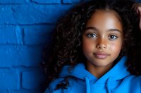 Young African American girl with natural curly hair wearing blue hoodie against vibrant blue brick wall background, expressing confidence and joy in casual portrait.