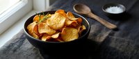 Golden crispy potato chips in black cast iron skillet on rustic wooden table with sea salt and herbs for gourmet snacking and appetizer presentation.