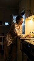 Middle-aged Caucasian woman in striped pajamas getting late night snack from refrigerator in modern kitchen with warm lighting and under-cabinet illumination.
