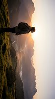 Hiker standing on mountain ridge at golden hour sunset with misty valley below, wearing dark jacket and backpack, enjoying scenic landscape view.
