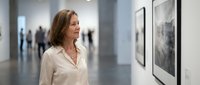 Middle-aged Caucasian woman in white blouse viewing framed artwork in modern gallery space with visitors in background for cultural exhibitions.