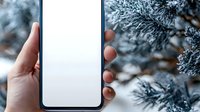 Hand holding modern smartphone with blank white screen against snowy winter pine branches background, mockup for app or website presentation.