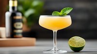 Golden cocktail in coupe glass with fresh mint garnish and lime half on marble counter with whiskey bottle background for bar menu design.
