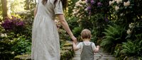 Mother and toddler son walking hand in hand through blooming garden path with colorful flowers and lush greenery creating peaceful family moment.