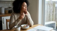 Young African American woman eating healthy breakfast bowl at kitchen table looking thoughtfully out window on rainy morning.