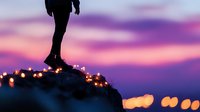 Silhouette of person standing on cliff edge against purple sunset sky and city lights bokeh, creating dramatic atmospheric scene of solitude and contemplation.