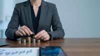 Professional businesswoman stacking coins at desk with financial charts, representing investment growth, savings strategy and wealth management planning.