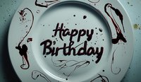 Artistic birthday message written in chocolate sauce on white plate with decorative swirls and splashes creating elegant calligraphy style lettering for celebration.