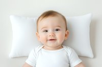 Smiling baby in white onesie lying on soft pillow against light background. Joyful expression captures pure innocence and happiness in minimalist setting.