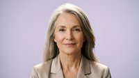 Mature woman with long gray hair and fair complexion wearing beige blazer against purple background, professional headshot for business, corporate, or executive use.