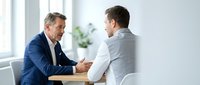Two male professionals in business attire engaged in discussion at wooden table in bright office setting with windows and plants. Suitable for corporate, recruitment, and business communication concepts.