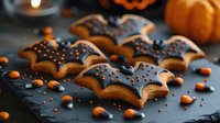Halloween bat-shaped cookies with chocolate glaze and orange sprinkles on dark slate surface with pumpkins and candy corn for autumn celebration.